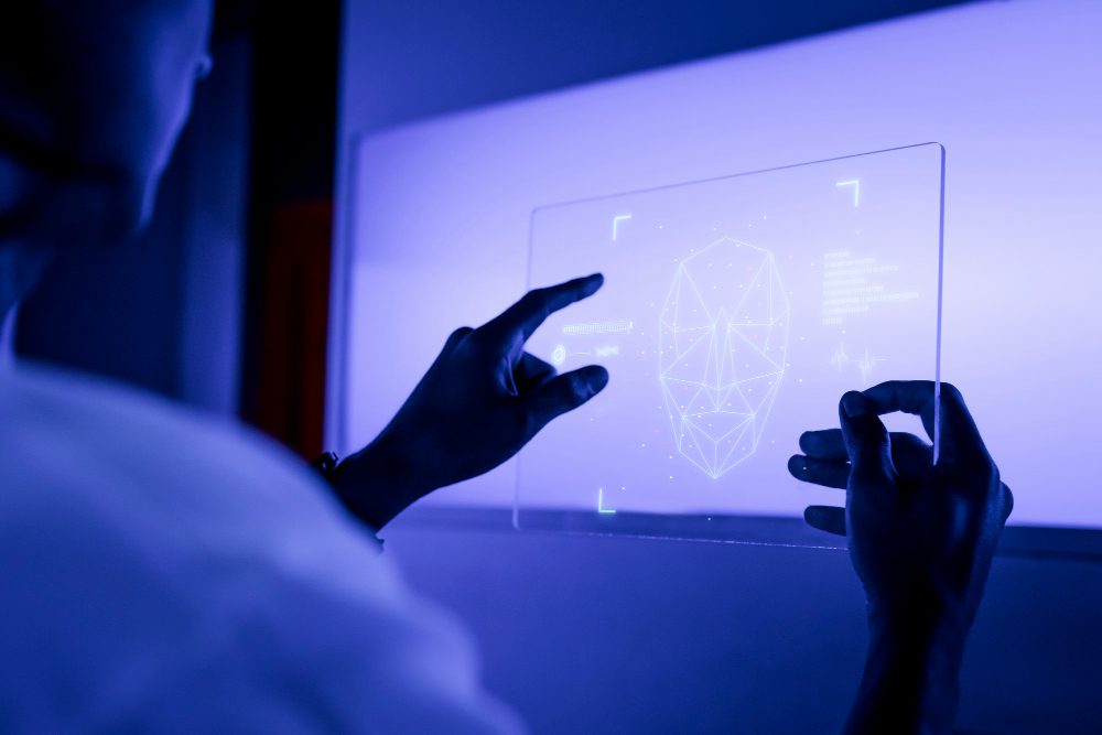Person uses a transparent touchscreen to manipulate a holographic wireframe of a human head on a blue-lit display.