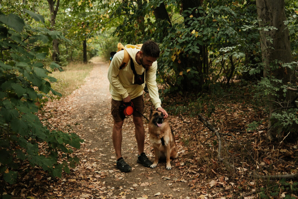 Man in a light hoodie pets a dog on a leaf-strewn forest path with a backpacked shoulder strap visible on his back.