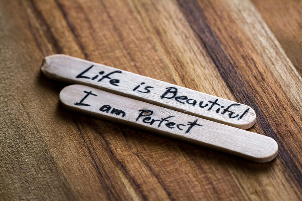 Two wooden popsicle sticks with handwritten motivational phrases: “Life is Beautiful” and “I am Perfect” on a wood surface, inspiring message pair.
