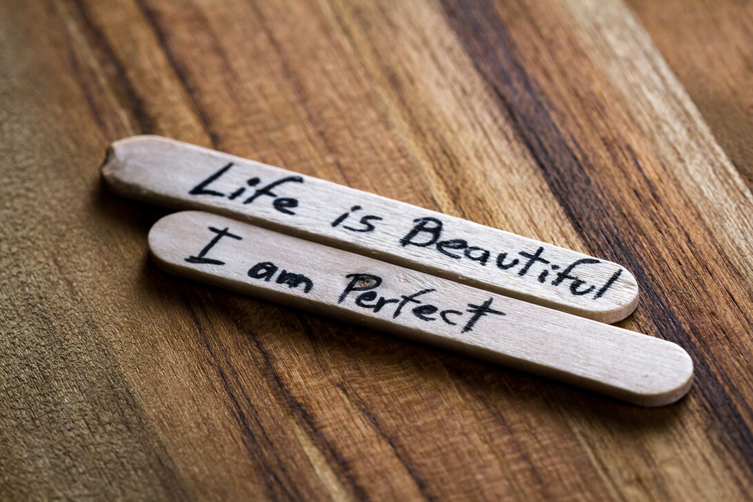 Two wooden popsicle sticks with handwritten motivational phrases: “Life is Beautiful” and “I am Perfect” on a wood surface, inspiring message pair.