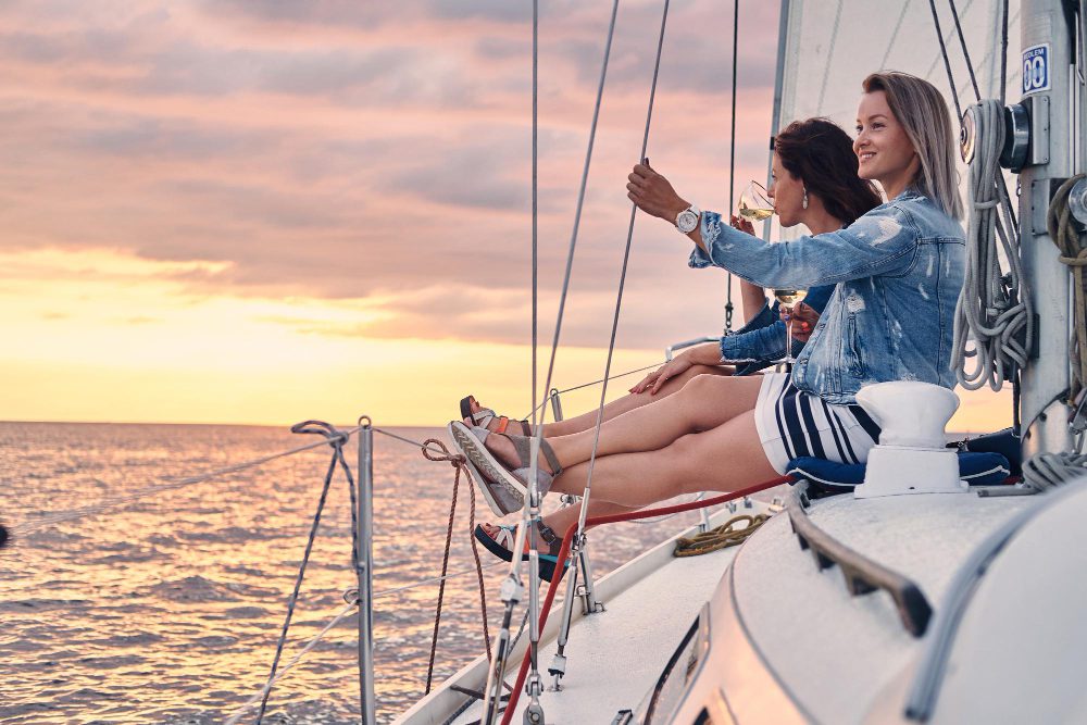 775800 - Ola120 - Europa Love 24h Three women sit on the edge of a sailboat at sunset, legs dangling, toasting with wine glasses on the deck