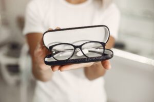 Black-rimmed eyeglasses held in an open black case toward the camera by a person in a white shirt.