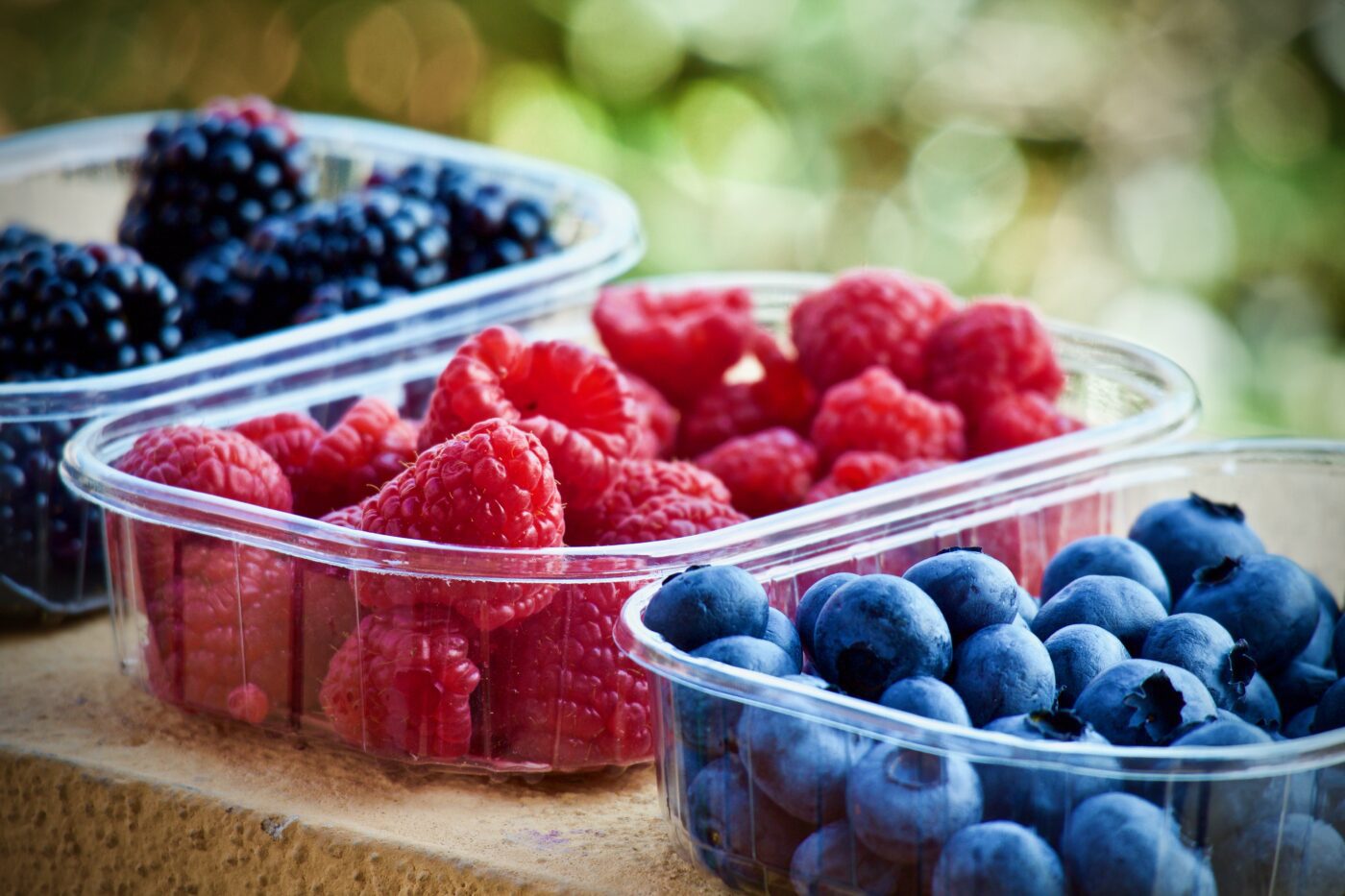 Fresh berries in clear containers: raspberries, blueberries, and blackberries on a stone surface outdoors.