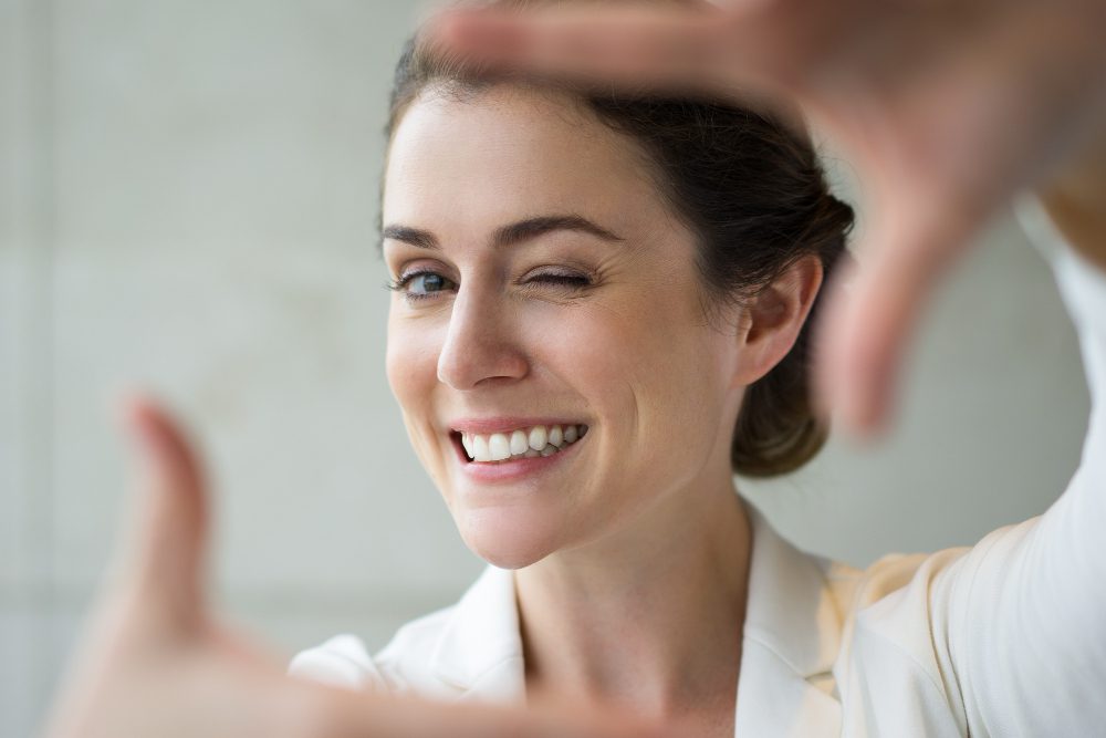 Woman smiling and winking at the camera while framing her face with her hands in front of a light background