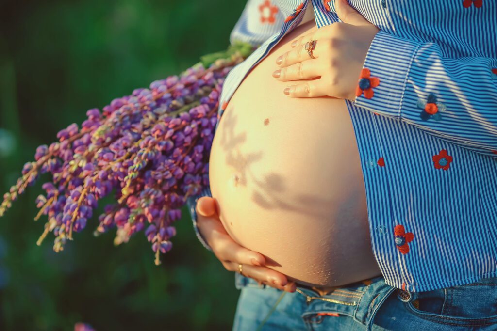 Pregnant woman's belly cradled by her hands outdoors, with a purple flower bouquet nearby.