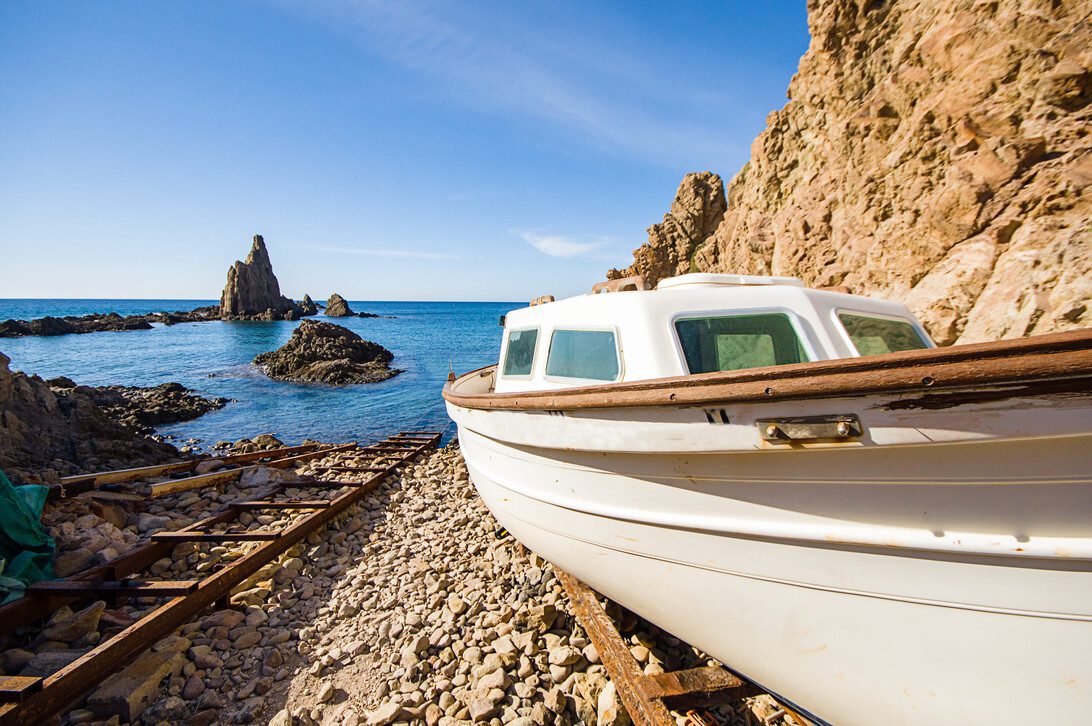 Cabo de Gata in Almeria Spain - Europa Love 24h White boat with wooden trim rests on a rocky pebble beach beside old rusted tracks, blue sea and cliff in the background.