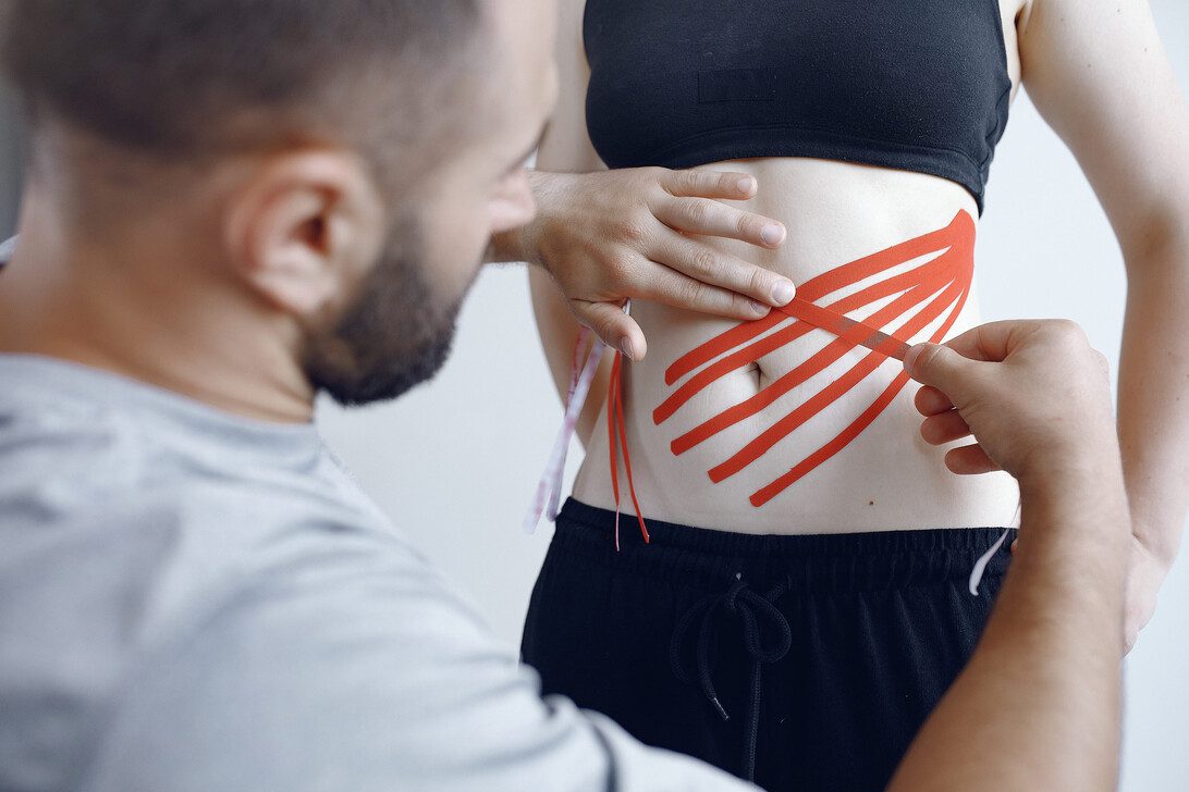Therapist applies red kinesiology tape in a crisscross pattern on a person’s midsection for support.
