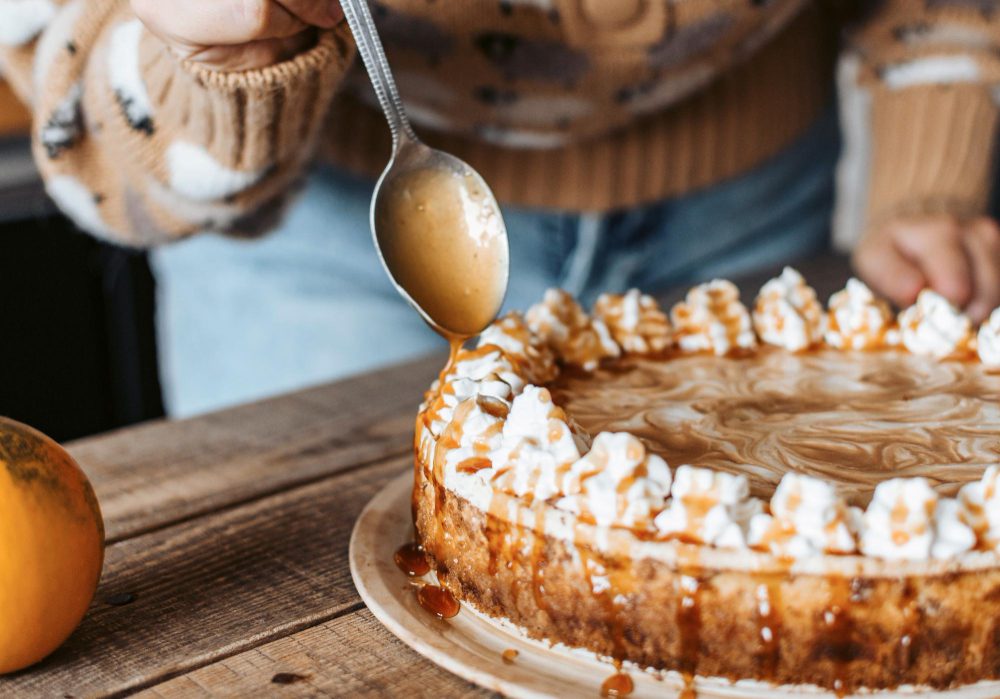 778458 - Longinos - Europa Love 24h Person in a cozy sweater drizzling caramel sauce over a caramel cheesecake edged with whipped cream on a rustic wooden table.