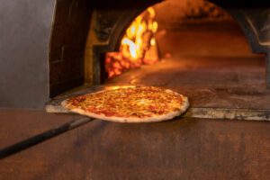 Pizza on a wooden peel sliding into a hot brick oven with blazing flames in the background, ready to bake.