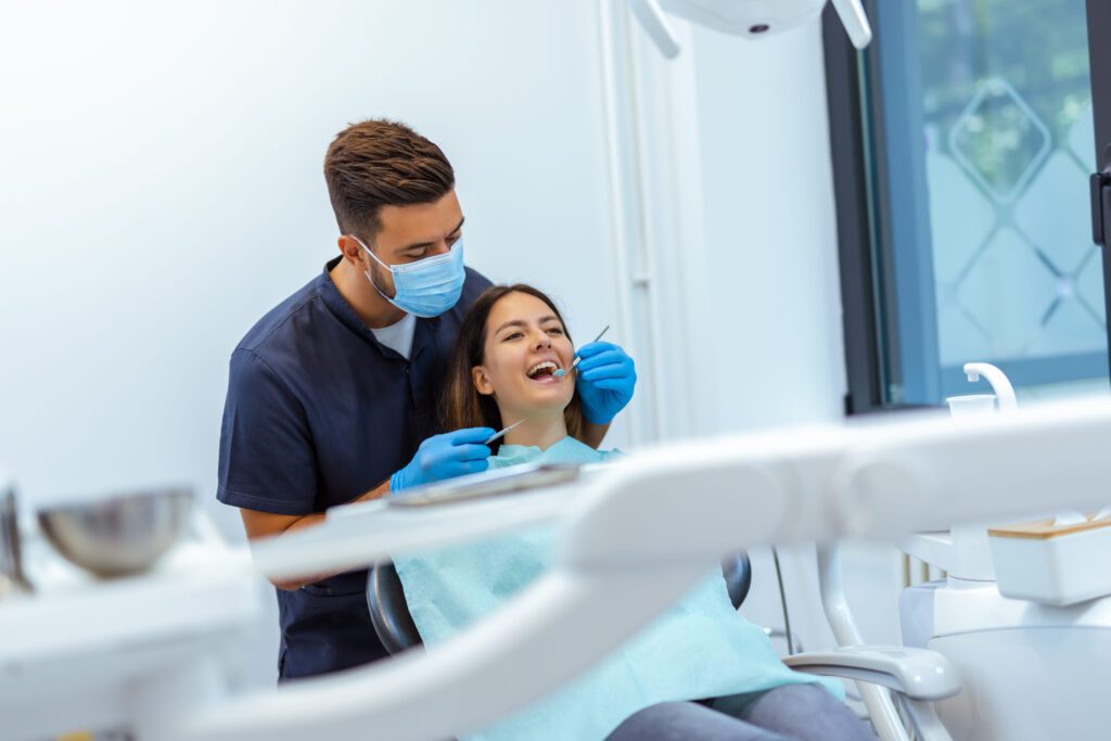 Dentist wearing a mask and gloves leans over a patient in a dental chair during a tooth cleaning or checkup.