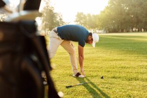 Golfer in a blue shirt and white cap bends to place a ball on the tee on a sunny golf course.