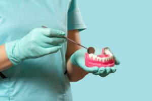 Dental professional in teal scrubs and gloves holds a pink dental model of teeth, examining it with a mirror tool against a turquoise background.
