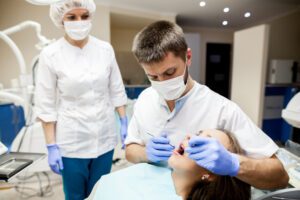 Dentist and dental assistant in masks and gloves treating a patient lying in a dental chair.