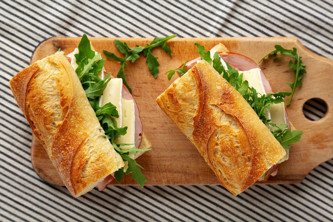Two crusty baguette sandwiches filled with cheese, ham and arugula on a wooden cutting board.